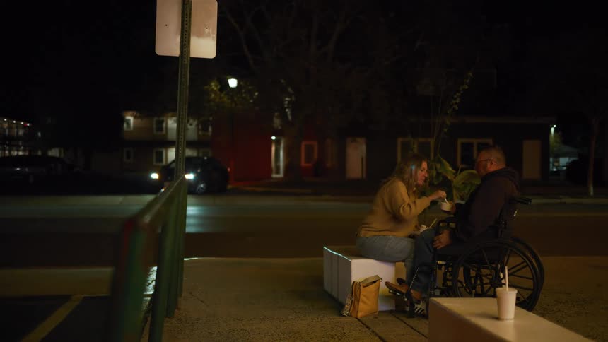 A woman sitting on a white box next to a man in a wheelchair on a dark sidewalk is seen sharing a casual meal with him offering him food from a container before they both eat illuminated primarily by streetlights and the headlights of passing cars