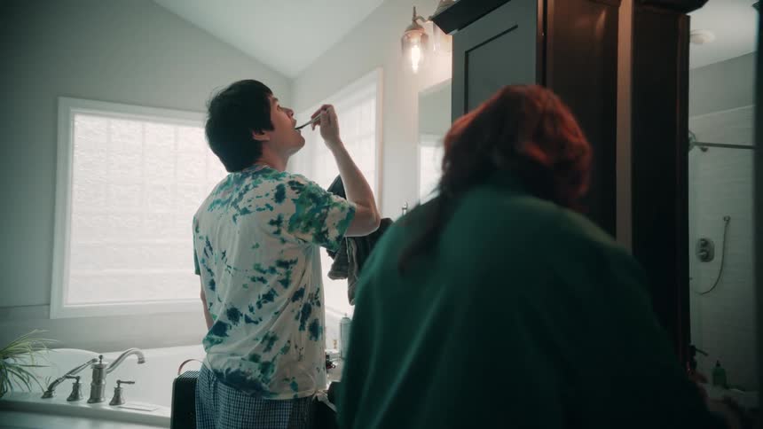 A young couple is inside the bathroom during daylight the man is brushing his teeth while the woman applies makeup both facing the mirror