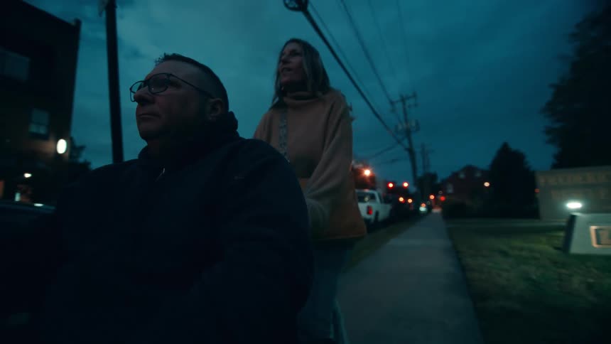 Adult woman pushing an adult man wearing glasses on a wheelchair on the sidewalk at an urban area outdoors during night time
