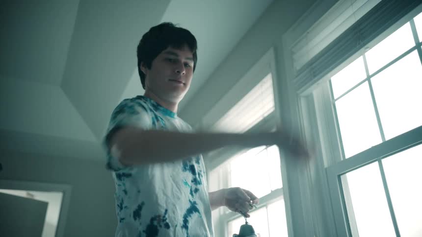 A young man in a blue and white tie dye shirt takes a striped shirt off a hanger near a brightly lit window in a dimly lit room