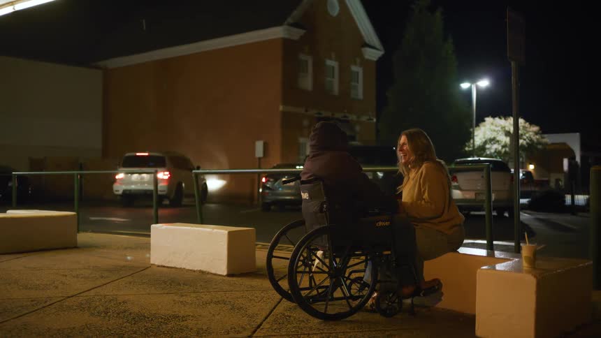 A woman sits on a low wall next to a man in a wheelchair and holds his hand talking and laughing under a streetlight at night in a parking lot