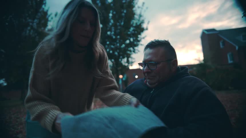 Woman covers a seated man with a blanket outdoors at dusk