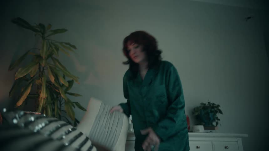 A woman with brown hair and bangs wears green silk pajamas as she picks up a pillow and makes the bed in a dimly lit bedroom