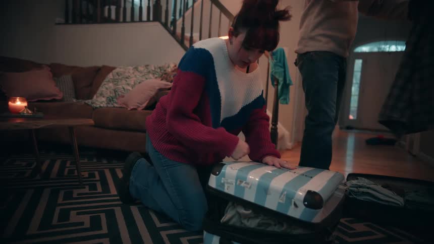 A woman forcefully closes a suitcase on the floor while a young man beside her folds clothes into another suitcase in the living room