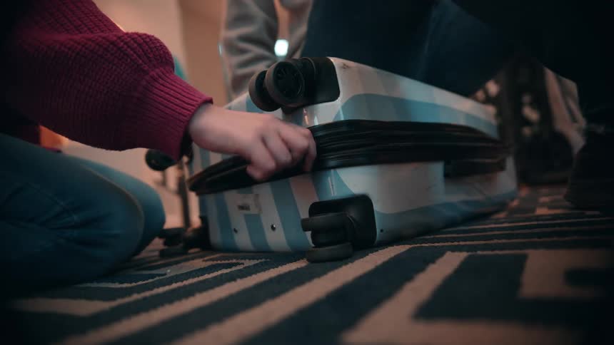 Two people are inside a room lit by a table lamp trying to open a briefcase placed on the floor