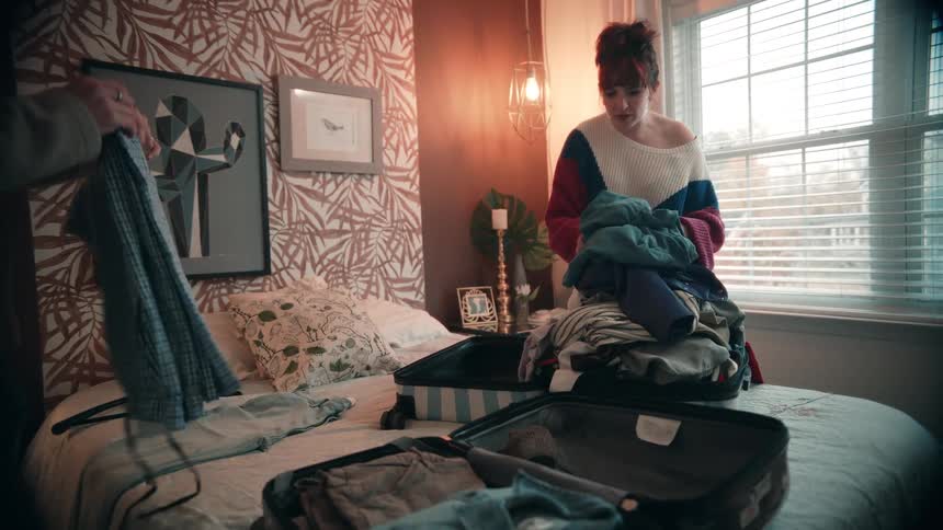 A young woman wearing a sweater stands at the foot of a bed in a decorated bedroom pushing clothes into a large suitcase overflowing with garments while a man joins her to help fold and place items into the open luggage