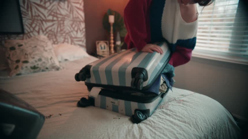 A woman packs clothes into a suitcase on a bed and leans on it to force it shut