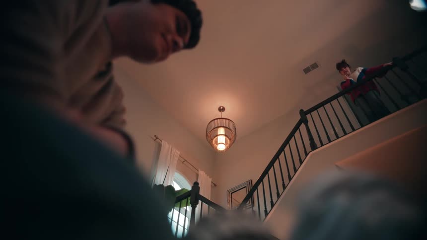 This short video clip shows a scene inside a home looking up from what appears to be a lower floor toward a staircase and a landingthere is a person in the foreground out of focus and another person standing on the landing in the background