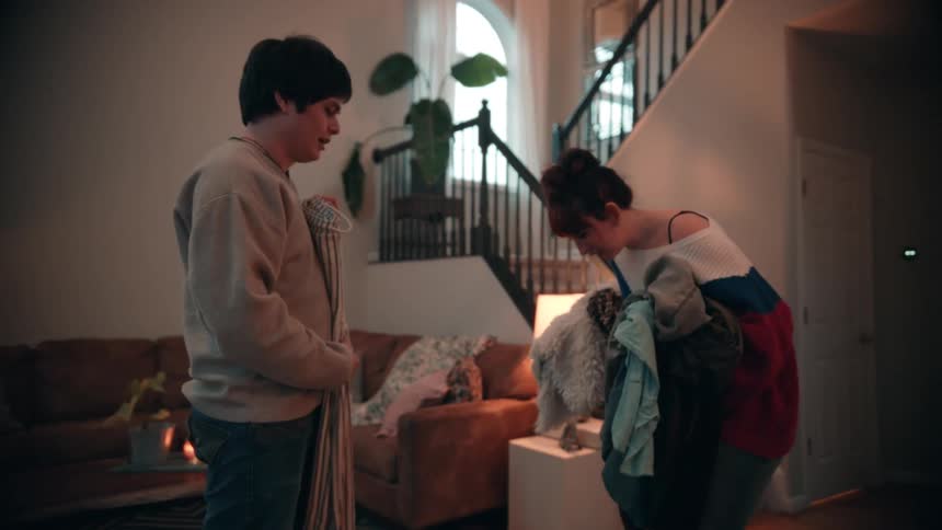 The video shows a man and a woman in the living area of a home standing near a staircase the woman initially descends the stairs carrying an armful of clothes and a bag while the man holds a striped piece of fabricthey meet at the bottom where they
