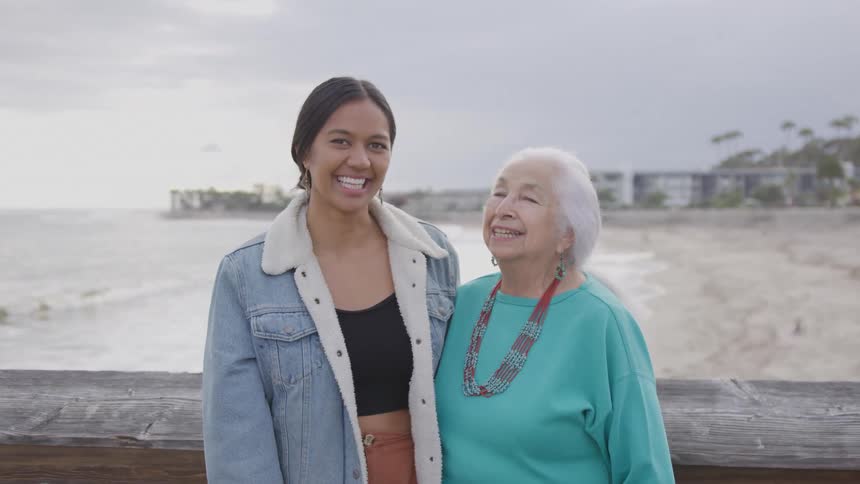 This short video clip features two women standing sidebyside on what appears to be a wooden pier or boardwalk with a beach and ocean in the backgroundthe younger woman on the left is smiling and looking directly at the camera she has long dark hai