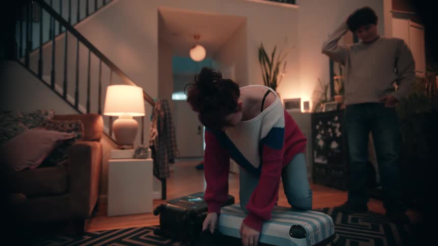 The scene takes place in a dimly lit living room or hallway where a woman is kneeling on a suitcase struggling to close it while packing a man stands nearby watching and running his hand through his hair suggesting a moment of stress or frustration