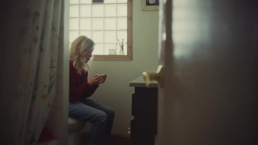 A sideprofile shot of a woman in a red sweater and jeans sitting on a toilet in a small bathroom intently examining a pregnancy test in her hands while waiting for a result