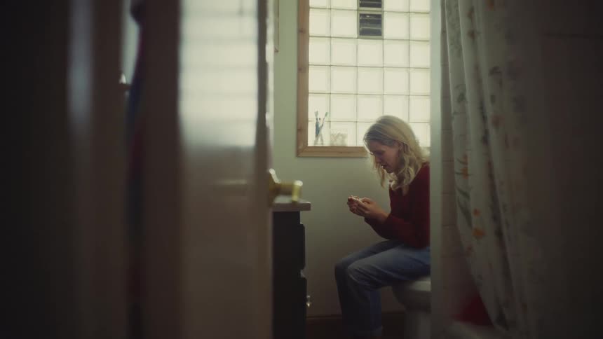 A blonde woman sits on a toilet in a bathroom looking anxiously at a pregnancy test in her hands next to a window with frosted glass blocks