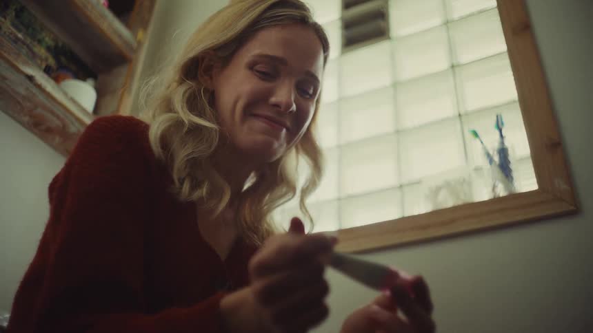 A woman with blonde hair seen from a low perspective holds a white test stick close to her face displaying a range of emotions from apprehension to a smile as she looks intently at the device in her hands inside a bathroom