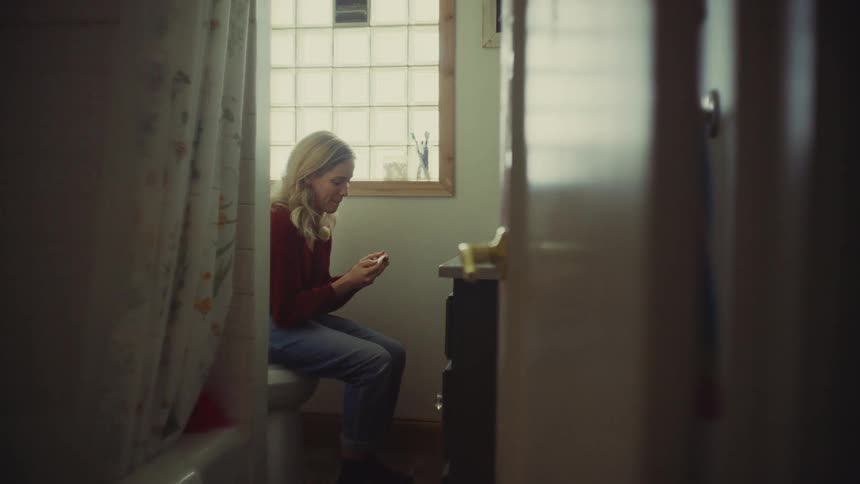 A young woman sits on the toilet at midday looking sadly at a pregnancy test tube in her hand