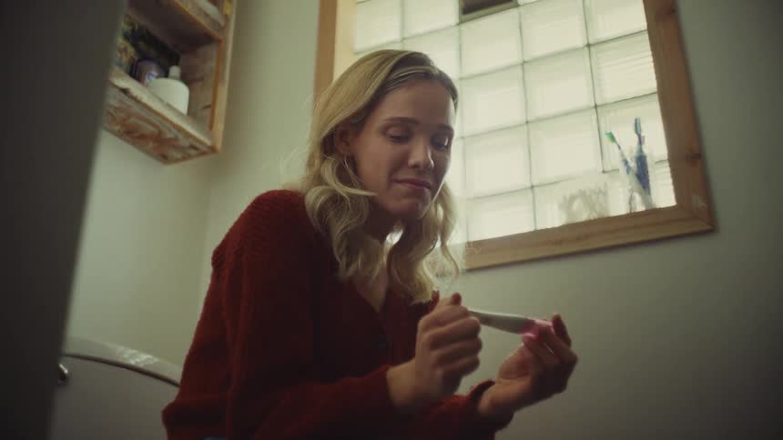 A young woman with blonde hair seated in a dimly lit bathroom holds a small white and pink pregnancy test device in her hands and exhibits an emotional expression of happiness looking to the result