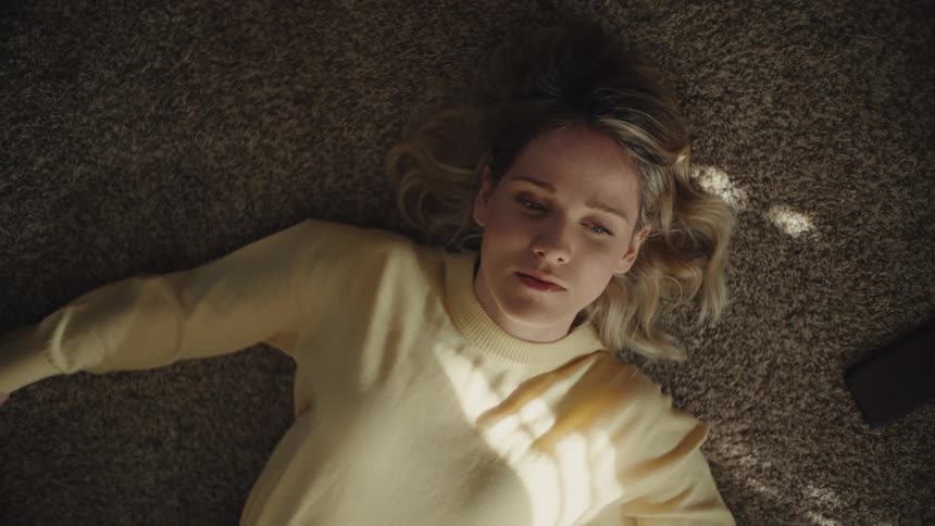 A blonde woman in a yellow sweater is lying on a brown textured carpet slowly opening her eyes to look around with a dazed expression before turning her head slightly