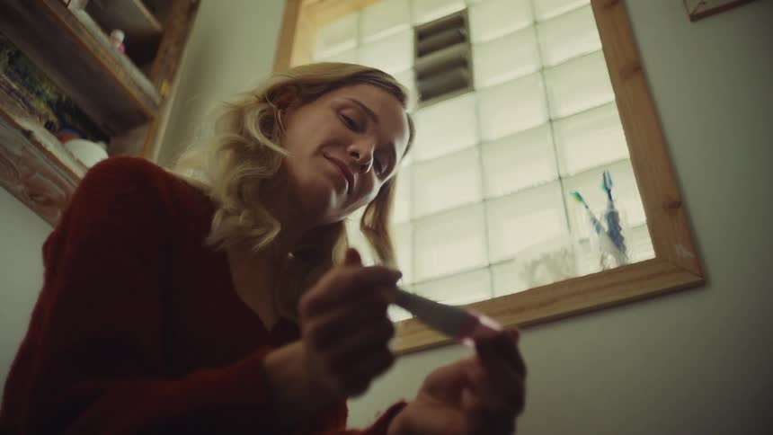 A blonde woman in a red sweater anxiously holds a pregnancy test looking up with a worried expression in a bathroom with a window and a mirror
