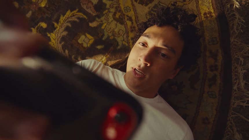 An overhead shot captures a man with curly hair lying on a patterned rug holding a mobile phone to film himself as he smiles and laughs into the camera lens