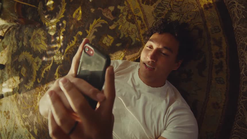 The man is texting on his cell phone while laying on a rug