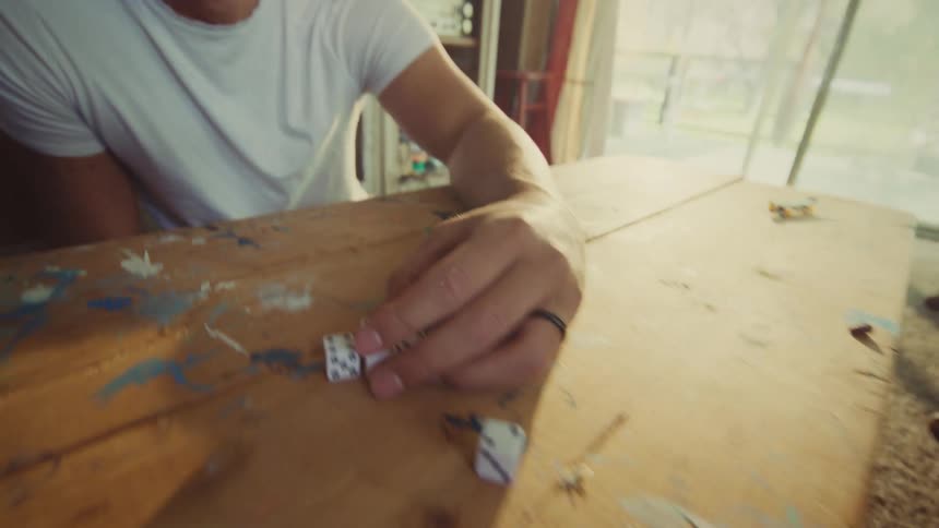 A smiling young man in a white t shirt throws a pair of dice onto a wooden table in a sunlit room with wood paneling