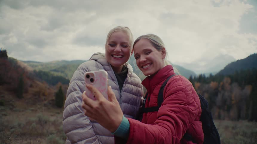 Two blonde women wearing puffer jackets are hiking outdoors and looking at a smartphone held up to take a selfie or view photosthey are smiling brightly at the phone against a backdrop of mountains covered in pine trees and a cloudy sky
