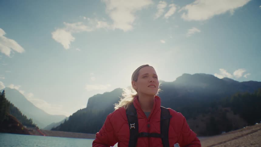 A woman with a backpack is standing next to a lake in the mountains