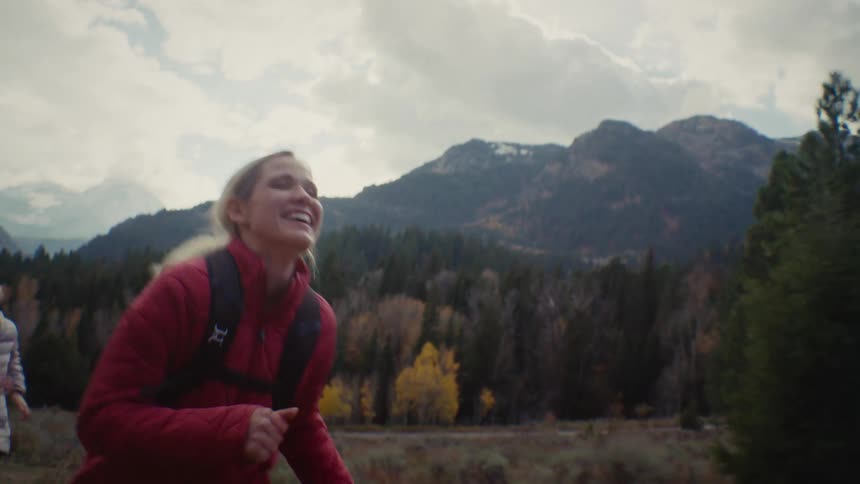 Two young adult woman running and laughing on a natural area surrounded by forests and mountains outdoors during day time