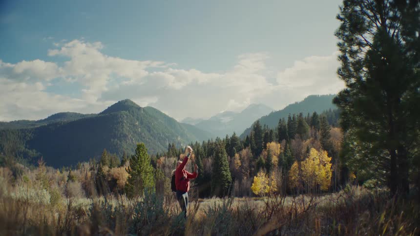 A woman is standing among trees in a forest on a mountain taking pictures with her phone