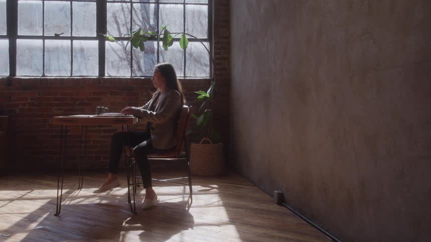 Woman walks into a room and puts her laptop computer on the desk as she opens the lid and sits down to type on the keyboard