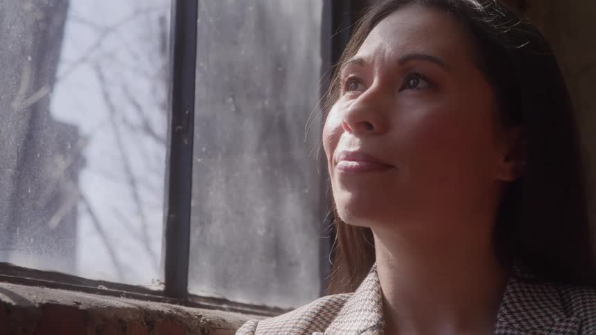 A thoughtful woman in a professional checkered jacket looks up at a bright industrial window with natural light hitting her face