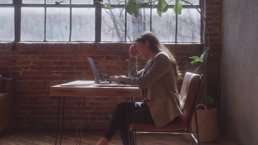 A stressed woman sits at a wooden desk with a laptop in a sunlit industrial office