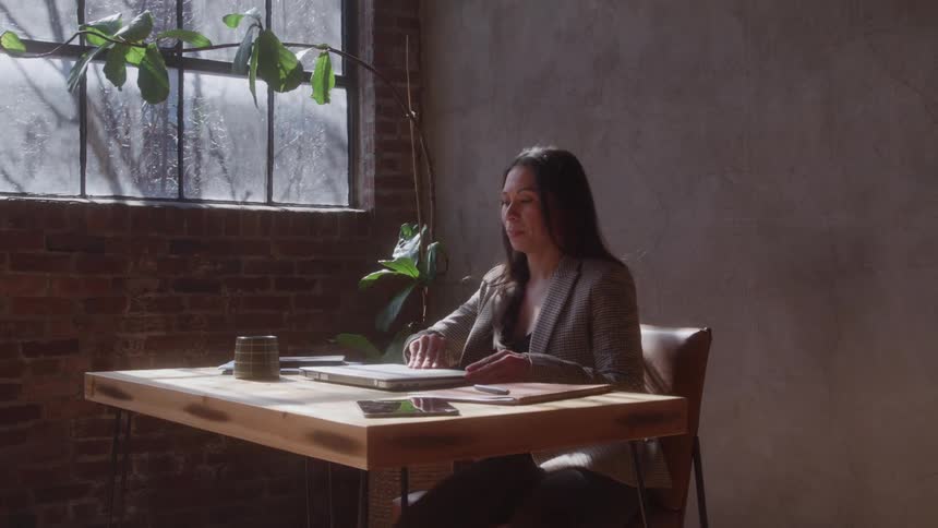 A young woman walks into a room sits behind a desk opens the lid of her laptop and begins typing while dressed in a suit