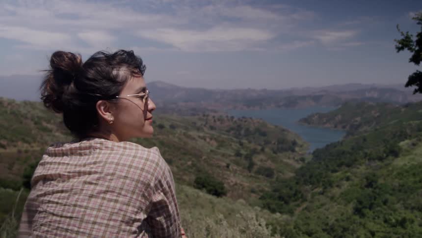 A woman is standing in the mountains looking down at a river