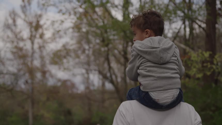A young child in a gray hooded sweatshirt sits on a mans shoulders looking forward with his back to the camera against a blurred outdoor background