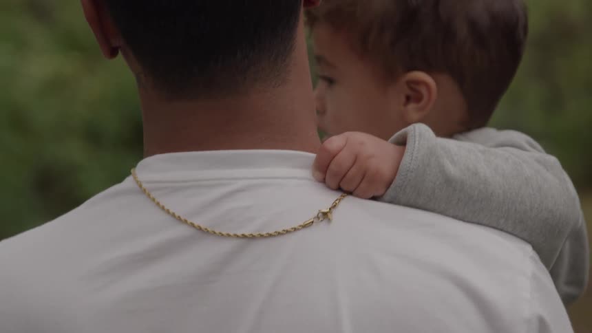 A close view from the back of a man wearing a gold chain and white shirt holding a child with curly hair over his shoulder outdoors