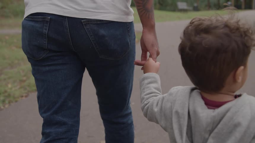 A rearview shot of an adult wearing a white tshirt and blue jeans and a small child in a gray hoodie walking sidebyside along a paved path in a park with the adult holding the childs hand