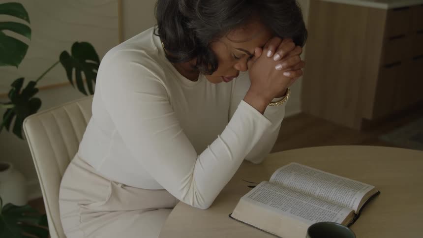 A woman in a white top sits at a table with an open book resting her head on her clasped hands in a moment of prayer or contemplation