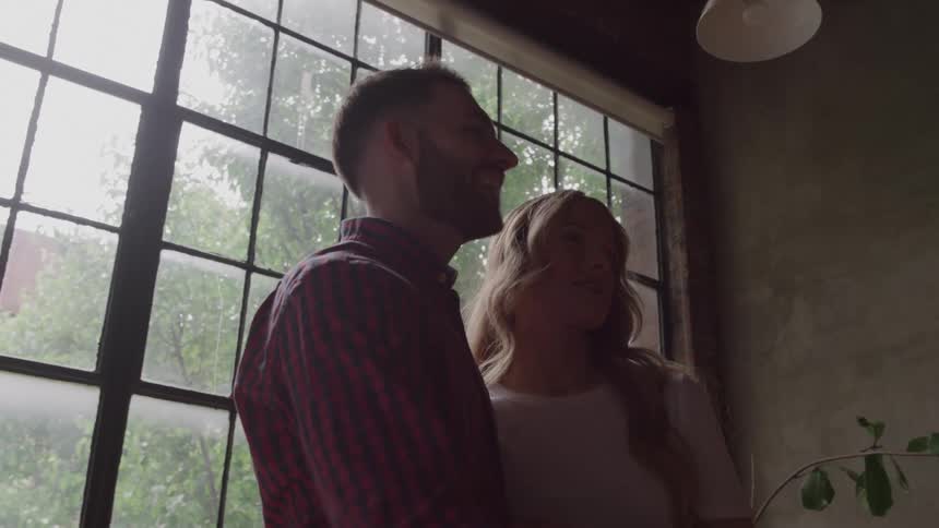 The video shows a man and a woman standing close together in an indoor space with a large industrialstyle window behind them the man has a beard and is wearing a red and black plaid shirt while the woman has long blonde hair and is wearing a white top
