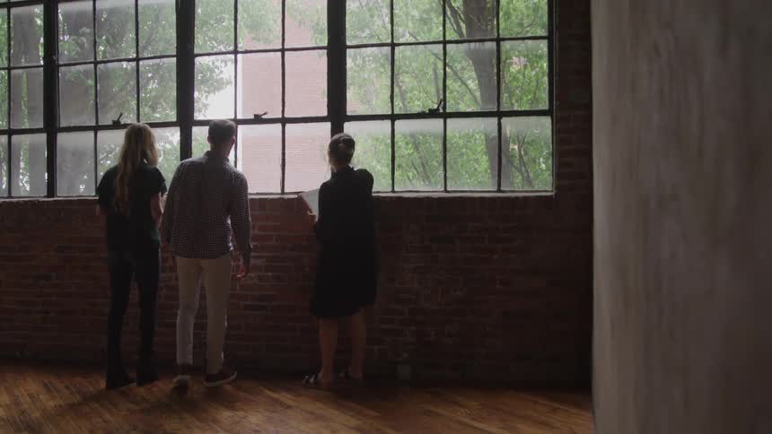 This video shows three people standing together looking out of a very large multipaned industrial window onto a leafy exteriorthey are in a spacious room with hardwood floors and an exposed brick wall possibly viewing a property for purchase or dis