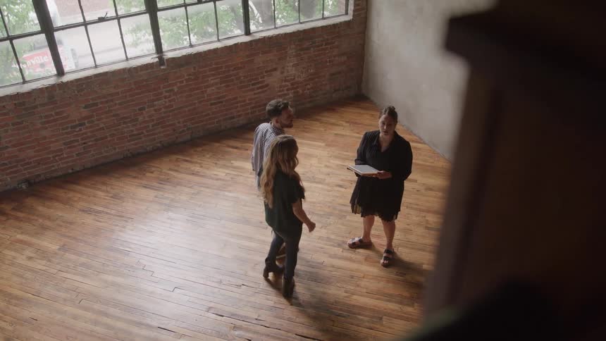 An overhead view of a realtor showing a large empty industrial style space with a brick wall hardwood floor and large windows to a man and a woman