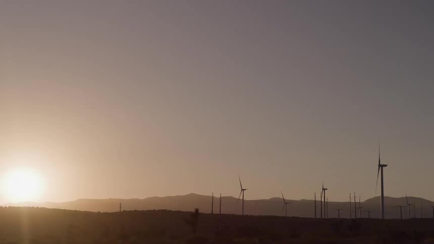 Turbines on a wind farm spin as the sun shines over mountains in the distance