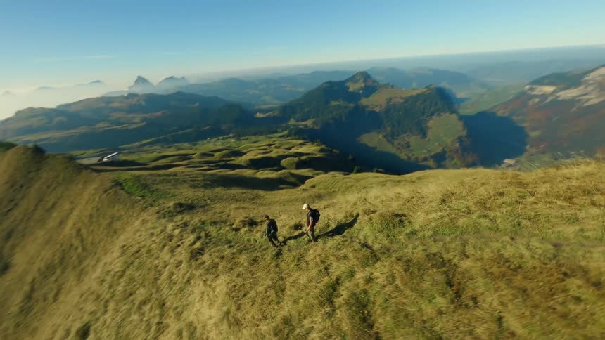 Two hikers walk down a grassy mountain ridge overlooking hills under bright sky