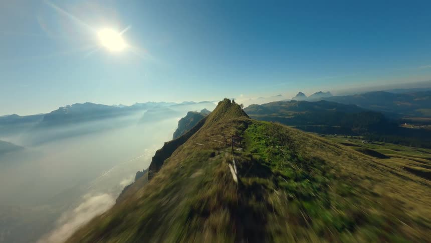 This aerial video clip features two hikers ascending and then standing on the narrow grassy ridge of a high mountain overlooking a vast landscapethe view showcases sharp mountain peaks some partially obscured by lowhanging fog or clouds in