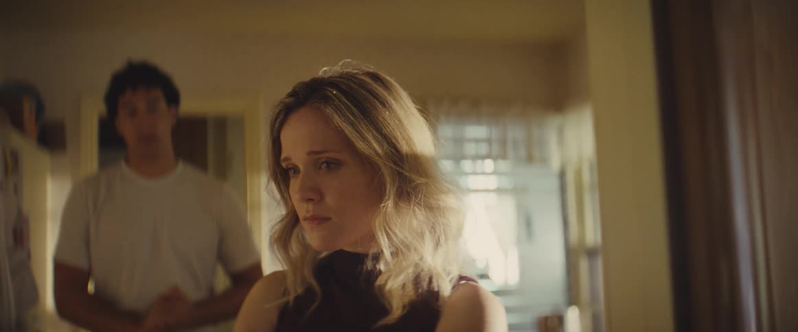 The video is a medium closeup of a blonde woman standing indoors looking off to the side with a serious or concerned expressiona man wearing a white shirt enters the background from the left suggesting a scene of interaction or tension between two