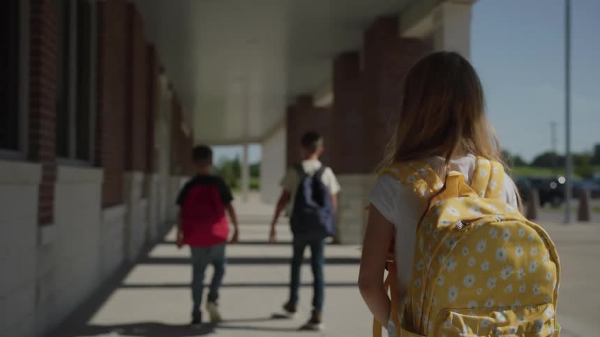 A girl wearing a bright yellow backpack with a white daisy pattern walks toward the entrance of a school building two boys with backpacks one red and one blue walk ahead of her under the shaded walkway on a sunny day
