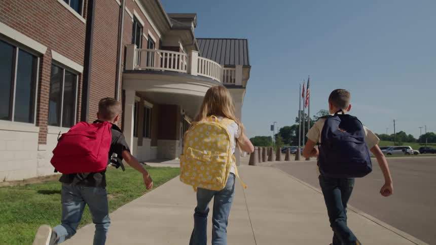 A camera follows three children two boys and a girl all wearing large backpacks as they run down a concrete sidewalk toward the main entrance of a brick school building on a sunny day