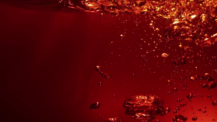 The video displays a closeup shot of numerous bubbles rising through a liquid that is intensely colored in deep red and orange huesthe movement of the bubbles suggests carbonation or fizzing in a beverage with the surface of the liquid visibly agitat