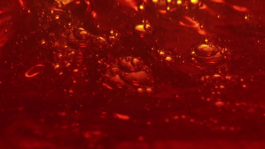 This is an extreme closeup of a viscous reddishamber liquid possibly syrup or oil bubbling and churning slowlythe movement highlights the fluids viscosity with small and large bubbles rising distorting the reflections of light within th