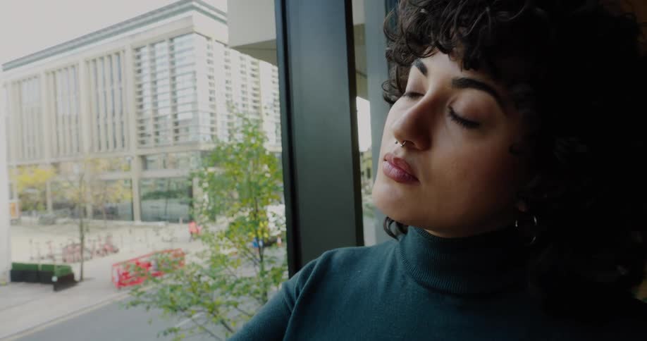 Young adult woman holding and looking at a handheld fan indoors next to a window showing a metropolitan area during day time